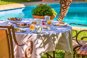 Frühstückstisch mit Orangensaft, Kaffee, Gebäck und Eiern neben türkisem Pool in Son Sant Jordi, Pollença. Sonnenlicht fällt durch Bäume.