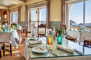 Elegantes Restaurantinterieur der Pousada Sagres mit Glastischen, buntem Glasgeschirr und Bogenfenstern mit Meerblick.
