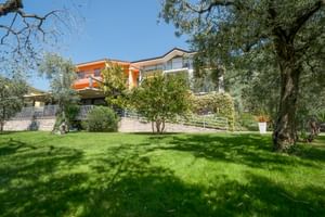Hotel Catullo in Malcesine mit orange-weißer Fassade, umgeben von grüner Rasenfläche, Olivenbäumen und mediterraner Vegetation unter blauem Himmel.