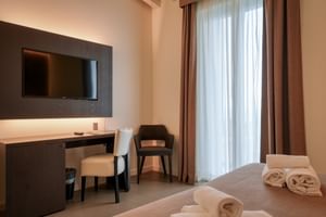 Modernes Hotelzimmer im Palazzo del Garda mit Wandfernseher, Schreibtischbereich, Stühlen und Bett mit weißen Handtüchern. Braune Vorhänge rahmen das Fenster.