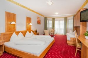 Spacious junior suite with wooden furniture, double bed with white linens, red patterned carpet, and three windows with curtains.