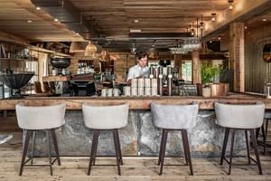 Modern alpine bar at Hotel Sepp in Maria Alm with wooden counter, white bar stools, coffee equipment, and bartender. Rustic wood ceiling.