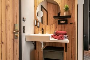 Modern bathroom with wood paneling, round mirror, white sink, black fixtures, and red towels at Hotel Astrid Saalbach.
