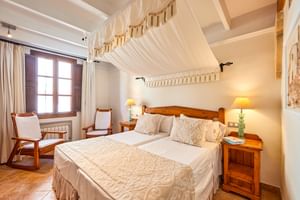 Helles Hotelzimmer in Pollença mit Doppelbett, weißem Himmelbett-Vorhang, Holzmöbeln, Terrakottaboden und zwei Nachttischlampen.