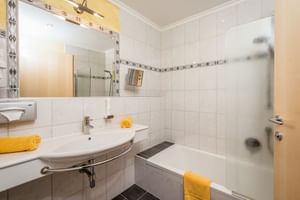 Modern bathroom with white tiles, sink with mirror and lighting, bathtub with glass partition, and yellow towel accents at Hotel Astrid Saalbach.