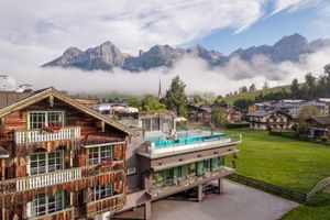 Hotel Sepp in Maria Alm with traditional wooden chalet and modern building with rooftop pool, surrounded by green meadows and Alpine peaks.