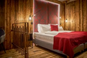 Modern hotel suite with white bed, red patterned headboard, wooden walls, and decorative wall lamps at Hotel Astrid Saalbach.