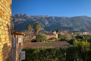 Blick vom Can Quatre in Sóller auf Steingebäude mit Terrakottadächern, Palmen und das Tramuntana-Gebirge unter klarem blauem Himmel.