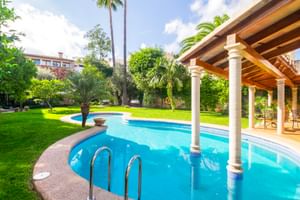 Geschwungener Pool mit weißen Säulen unter überdachter Terrasse, umgeben von saftigem Rasen und Palmen in Son Sant Jordi in Pollença.