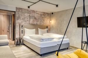 Modernes Doppelzimmer mit beigem Polsterbett, dekorativer Musterwand, Holzboden, gelbem Hängesessel und Schienenbeleuchtung.