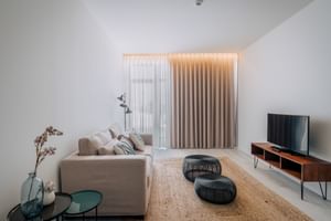 Helles Wohnzimmer mit beigem Sofa, Holz-TV-Möbel, Juteteppich, schwarzen Poufs und weißen Vorhängen. Minimalistisches Dekor mit Dachschräge.