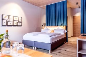Modern double room at Lechzeit Elmen with gray bed, blue curtains, wooden ceiling, and framed botanical prints on white walls.