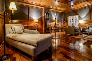 Elegante Lounge mit Holzvertäfelung, Kassettendecke, beiger Chaiselongue, Standuhr und Sitzbereichen mit warmer Beleuchtung.