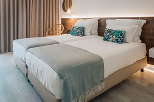 Modernes Doppelzimmer mit zwei Betten, weißer Bettwäsche, türkisfarbenen Kissen, Holzkopfteil, beigen Vorhängen und Korbstuhl im Praia Hotel Porto Covo.