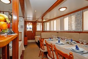 Dining area on MS Sagena with set tables, wooden chairs, beige upholstery, and warm wood paneling. Fruit basket visible on left counter.