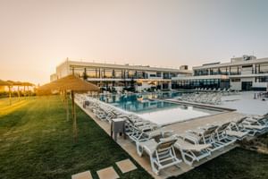 Außenpool im Praia Hotel & Spa Porto Covo mit weißen Sonnenliegen auf Rasen und modernem Hotelgebäude im Hintergrund bei Sonnenuntergang.