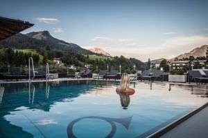 Frau im Infinity-Pool des Hotel Alte Post Fieberbrunn mit Bergpanorama. Sonnenliegen säumen das Pooldeck mit Alpengipfeln im Hintergrund.
