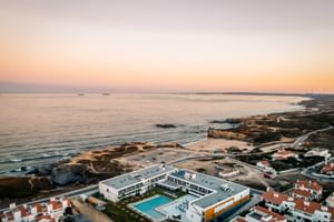 Luftaufnahme des Praia Hotel & Spa Porto Covo bei Sonnenuntergang mit weißem modernen Gebäude und Pool an felsiger Küste.