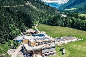 Aerial view of Lechzeit Elmen hotel with solar panels on roof, surrounded by green meadows and forested mountains in Austrian Alps.