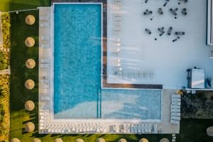 Luftaufnahme des Praia Hotel Porto Covo Pools mit weißen Sonnenliegen, Strohdächern auf Rasen und angrenzender Terrasse mit dunklen Sitzgelegenheiten.
