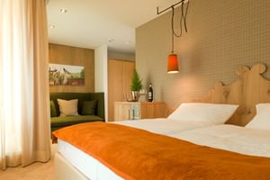 Modern hotel room with wooden headboard, orange bed runner, green seating area, orange pendant light and wooden furnishings.