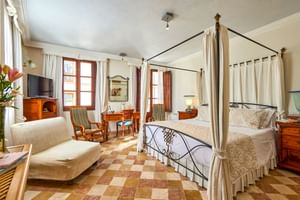 Geräumiges Doppelzimmer mit Himmelbett aus Eisen, kariertem Fliesenboden, Holzmöbeln, beigem Sofa und Fenstern mit weißen Vorhängen in Pollença.