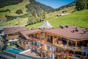 Aerial view of Hotel Astrid in Saalbach with wooden balconies and outdoor pool, surrounded by green Alpine meadows and ski slopes.