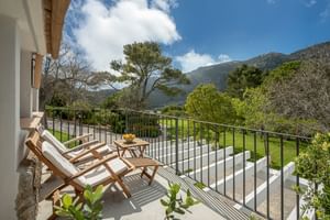 Sonniger Balkon im Son Palou Orient mit zwei Holzliegen und Tisch mit Blick auf grüne Gärten und Berge unter blauem Himmel mit Wolken.