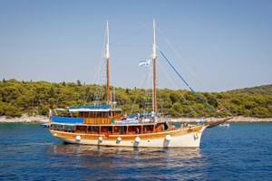 Wooden sailing ship MS Sagena with two masts and blue canopy anchored in calm blue water. Green forested coastline visible in background.