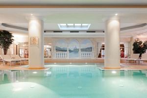 Elegant Tirolerhof indoor pool with white columns, turquoise water, and mountain landscape mural. Skylights illuminate the luxurious space.