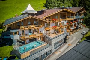 Aerial view of Hotel Astrid Saalbach, an alpine wooden hotel with balconies and an outdoor pool, surrounded by green meadows and forest.