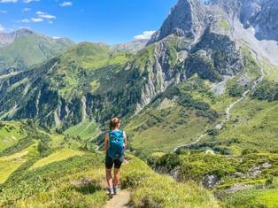 Hiker with blue backpack walking on mountain trail overlooking green valleys and rocky peaks in the Allgäu-Lechtal Alps on the Arlberg Trail.