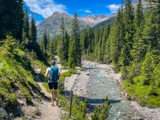 Hiker with backpack walking on trail beside mountain stream. Dense conifer forest lines both sides, with rocky peaks under blue sky.