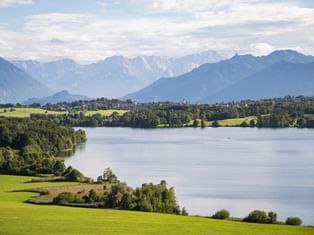 Scenic view of a lake surrounded by green meadows and forests with snow-capped Alpine mountains in the background under a cloudy sky.