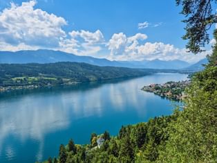 Panoramablick auf den Millstätter See von der Luschan Höhe Plattform. Blauer See umgeben von grünen Wäldern, kleine Dörfer am Ufer, Berge im Hintergrund.