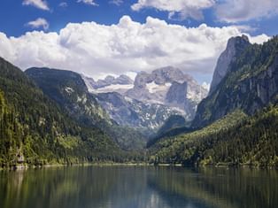 Gosausee lake in Salzkammergut with calm water reflecting forested mountains. Dachstein massif with snow-capped peaks visible in background.