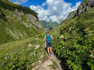 Hiker with blue backpack walking on mountain trail through green valley. Rocky cliffs on both sides, distant peaks and clouds visible.