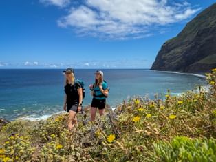 Two female hikers with backpacks standing on a coastal cliff in Madeira, surrounded by yellow wildflowers, overlooking the blue ocean.