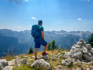 Hiker with backpack standing on rocky summit in the Julian Alps, overlooking mountain ranges under blue sky with stone cairn nearby.