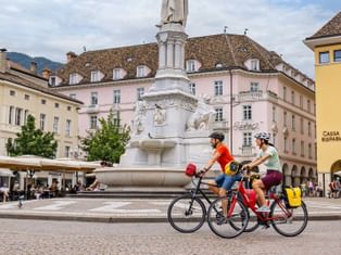 Two cyclists riding past the white monument fountain at Waltherplatz in Bolzano, with historic pastel-colored buildings and outdoor cafes.