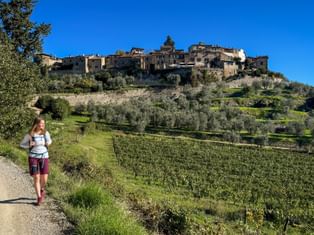 Female hiker with backpack walking on gravel path through green hillside with olive trees. Historic stone village visible on hilltop under blue sky.