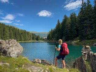 Female hiker with pink backpack and trekking poles standing by turquoise Obernberger See, surrounded by coniferous forests and mountains.