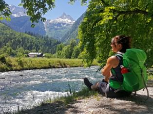 Wanderin mit grünem Rucksack sitzt an einem Gebirgsfluss und blickt auf die Loferer Steinberge. Bewaldete Hänge und Almwiesen umgeben die Szenerie.