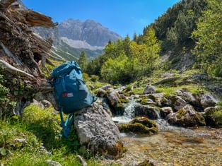 Teal backpack resting on a rock beside a mountain stream with moss-covered stones. Alpine peaks and green forest visible in the background.