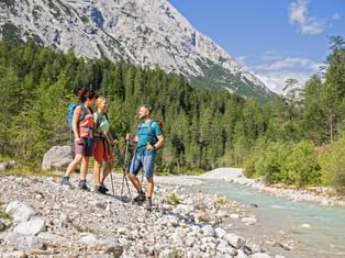 Three hikers with backpacks and trekking poles standing on rocky riverbank of Leutascher Ache in Bavaria, with mountain and forest backdrop.