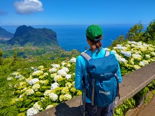 Female hiker with green cap and blue backpack standing among white hydrangea flowers, overlooking Madeira's dramatic coastline and blue ocean.