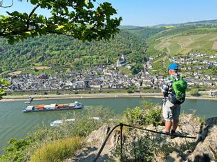 Hikers on the rock of the Loreley with a view of a village and cargo ships on the Rhine