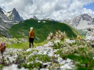 Hiker in orange jacket standing on alpine meadow with wildflowers, viewing Weissschrofenspitze peak and dramatic mountain landscape.