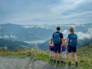 Family of four with backpacks viewing alpine panorama in Pinzgau. Mountain lake and peaks visible below through clouds.