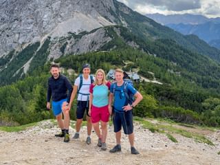 Vier Wanderer mit Rucksäcken auf dem Vršič-Pass-Gipfel mit bewaldeten Bergen und felsigen Gipfeln der Julischen Alpen im Hintergrund.